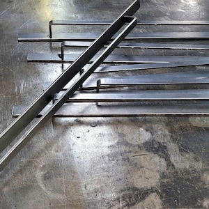 Pode incluir: Um conjunto de seis barras de metal com pontas arredondadas, dispostas em um padrão escalonado sobre uma superfície cinza. As barras são todas do mesmo comprimento e largura, e são feitas do mesmo tipo de metal.