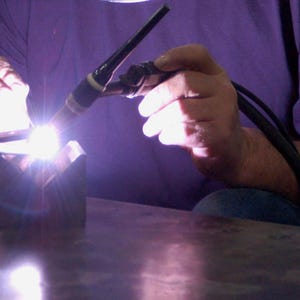 May include: A person welding metal with a torch, creating a bright, focused light. The welder is wearing a purple shirt. The metal is being joined together with a focused beam of light.
