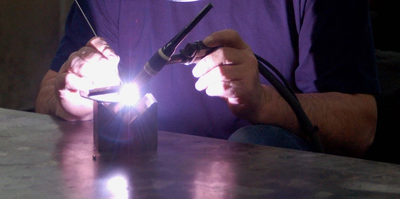 May include: A person welding metal with a torch, creating a bright, focused light. The welder is wearing a purple shirt. The metal is being joined together with a focused beam of light.
