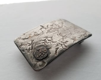 May include: A silver belt buckle with a textured surface and a small, circular design in the center.