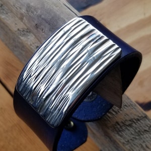 Op de afbeelding: Een donkerblauwe leren armband met een rechthoekige, getextureerde plaat van zilver.