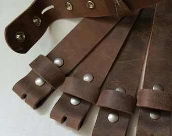 Puede incluir: Cinco correas de cuero marrón con hebillas de metal plateado. Las correas están dispuestas en forma de abanico, con el extremo de la hebilla de cada correa apuntando hacia el centro del abanico.