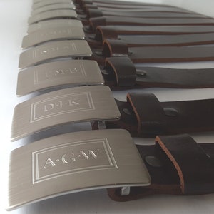 May include: A row of silver belt buckles with engraved initials. The buckles are attached to brown leather belts.
