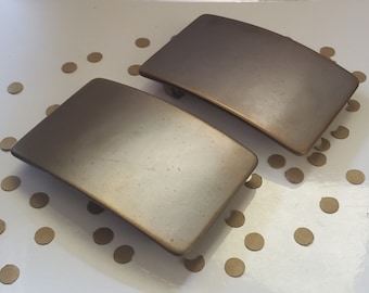 May include: Two gold-toned metal hair clips. The clips are rectangular and have a slightly curved shape. They are both made of a smooth, shiny metal.