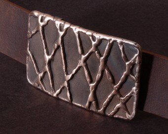 May include: A silver belt buckle with a textured, geometric design. The buckle is rectangular and has a slightly rounded edge.