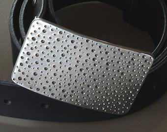 May include: A silver-colored rectangular belt buckle with a pattern of small, evenly spaced holes. The buckle is set against a black leather belt, with the buckle's surface reflecting light. The design is modern and minimalist.