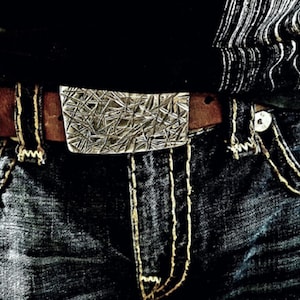 Peut inclure: Une ceinture en cuir marron avec une boucle argentée ornée d'un motif géométrique. La ceinture est portée sur un jean bleu avec des coutures jaunes.