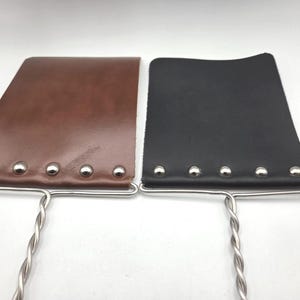 May include: Two rectangular leather paddles, one brown and one black, each featuring five silver-toned metal rivets along the bottom edge. The leather pieces are attached to twisted silver-toned metal handles that extend downwards from the base.