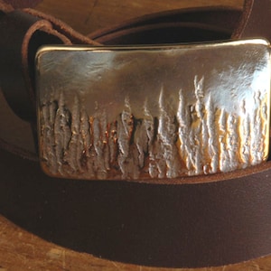 May include: A brown leather belt with a silver buckle featuring a textured design of trees.