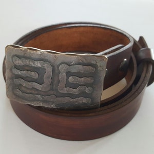 May include: A brown leather belt with a rectangular, textured metal buckle. The buckle has a dark, etched design. The belt is rolled up, showing the buckle and the leather strap.