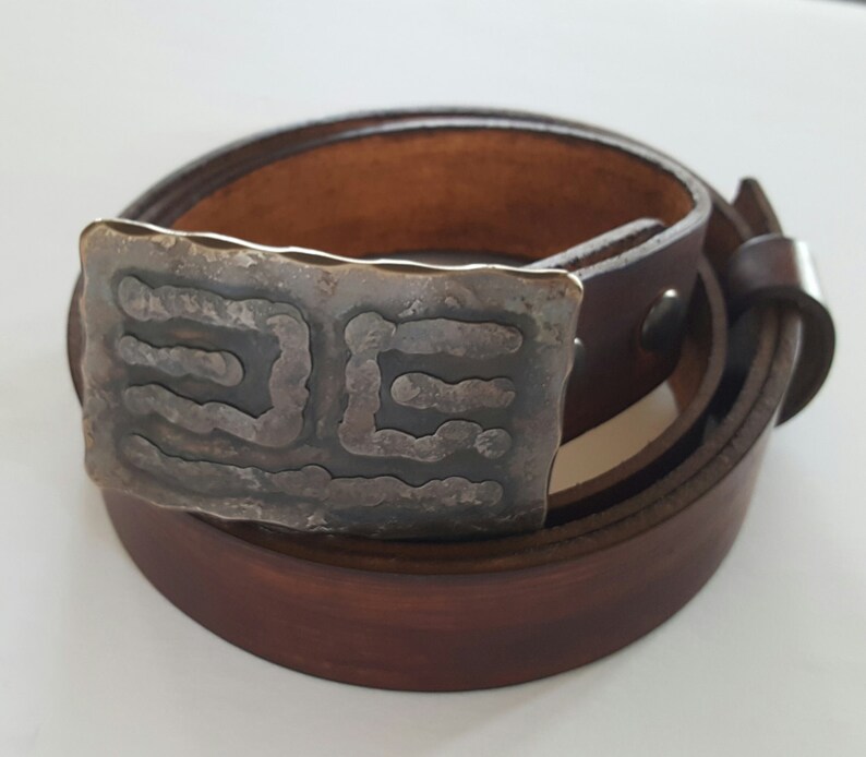 May include: A brown leather belt with a rectangular, textured metal buckle. The buckle has a dark, etched design. The belt is rolled up, showing the buckle and the leather strap.