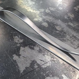 May include: A silver metal handle with a curved design. The handle is on a flat, silver metal surface.