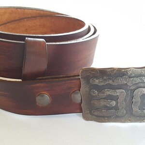 May include: A brown leather belt with a decorative metal buckle. The belt is rolled up, showing the inside and the buckle is rectangular with a textured design. The belt is a rich brown color.