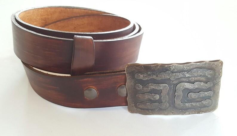 May include: A brown leather belt with a decorative metal buckle. The belt is rolled up, showing the inside and the buckle is rectangular with a textured design. The belt is a rich brown color.