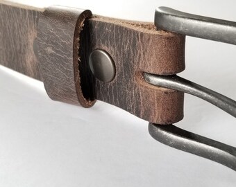 May include: Close-up of a brown leather belt with a silver-colored buckle. The belt is made of textured, distressed leather and features a metal buckle and a metal snap closure. The belt is approximately 1.5 inches wide.