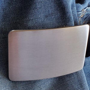 May include: A silver metal belt buckle with a smooth, brushed finish. The buckle is attached to a black leather belt.