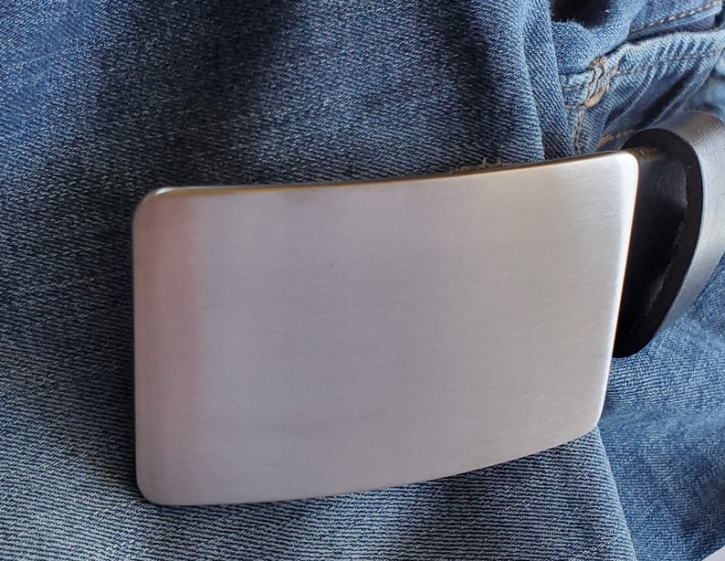 May include: A silver metal belt buckle with a smooth, brushed finish. The buckle is attached to a black leather belt.