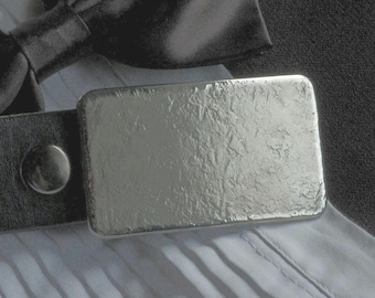 May include: A silver belt buckle with a textured surface. The buckle is attached to a black leather belt.