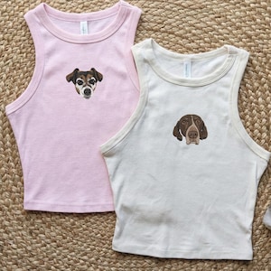 Puede incluir: Dos camisetas sin mangas, una rosa claro y otra blanquecina, cada una con una cara de perro bordada. La camiseta rosa tiene un perro marrón y blanco, mientras que la blanquecina tiene un perro marrón y blanco. Sobre una estera tejida.