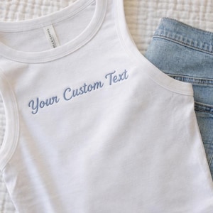 May include: White tank top with the embroidered text "Your Custom Text" in a light blue script. The tank top is paired with light blue denim jeans. The jeans have a brown leather patch on the back.