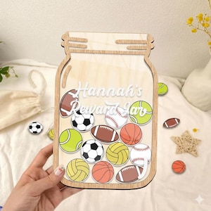 May include: Wooden jar-shaped reward chart with the text "Hannah's Reward Jar". The jar contains illustrations of various sports balls, including soccer, baseball, and football. The jar is made of light-colored wood.