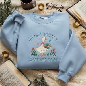 Può includere: Felpa azzurra con ricamo. Il disegno presenta un'oca bianca che tiene dei libri, circondata da fiori, e la scritta "OOPS, I BOUGHT ANOTHER BOOK". La felpa ha girocollo e maniche lunghe.