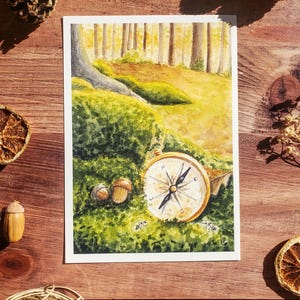 May include: A rectangular watercolour print featuring a forest scene with a gold compass resting on a mossy patch next to two acorns. The background shows tall tree trunks and a yellow-toned path. The compass face displays cardinal directions.