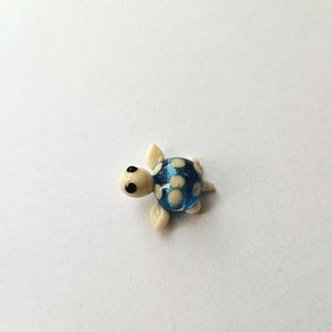 May include: A small, handcrafted glass turtle figurine. The turtle has a blue shell with white polka dots, a cream-coloured head, and four cream-coloured flipper-like appendages. The eyes are black. The figurine is a decorative item.