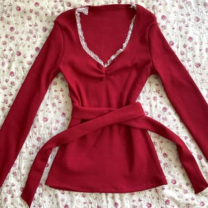 May include: A long-sleeved, red top with a V-neckline trimmed with white lace. The top features a cinched waist and a matching tie belt. The fabric has a ribbed texture, and the overall style is feminine and classic.
