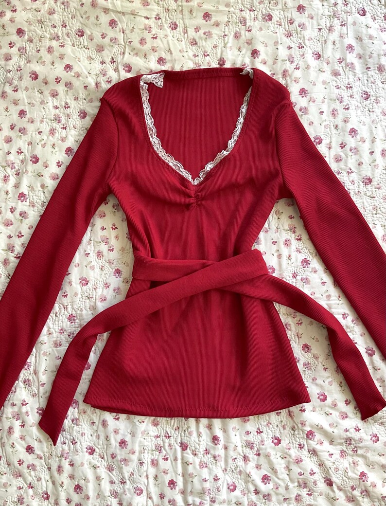 May include: A long-sleeved, red top with a V-neckline trimmed with white lace. The top features a cinched waist and a matching tie belt. The fabric has a ribbed texture, and the overall style is feminine and classic.