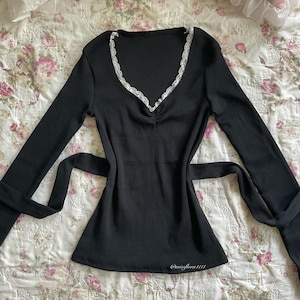 May include: A black long-sleeve top with a V-neckline trimmed with white lace. The top has a ribbed texture and a tie at the waist. The text "@missflora1111" is visible near the bottom hem.