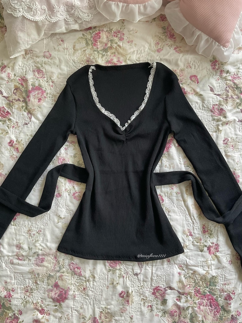 May include: A black long-sleeve top with a V-neckline trimmed with white lace. The top has a ribbed texture and a tie at the waist. The text "@missflora1111" is visible near the bottom hem.