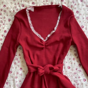 May include: A long-sleeved, red dress with a V-neckline trimmed with white lace. The dress has a cinched waist with a matching fabric belt tied in a bow. The fabric has a ribbed texture.