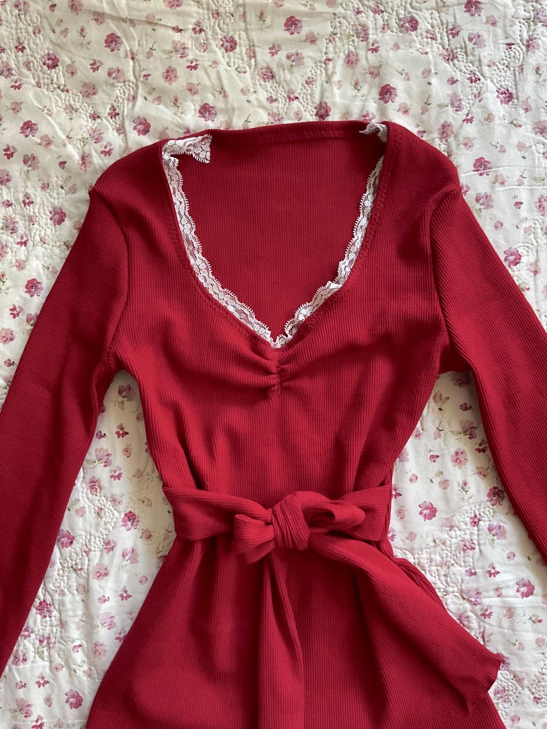 May include: A long-sleeved, red dress with a V-neckline trimmed with white lace. The dress has a cinched waist with a matching fabric belt tied in a bow. The fabric has a ribbed texture.