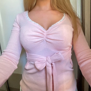 May include: A light pink long-sleeved top with a V-neckline and a bow tie belt. The neckline is trimmed with white lace. The top has a ruched detail at the bust.
