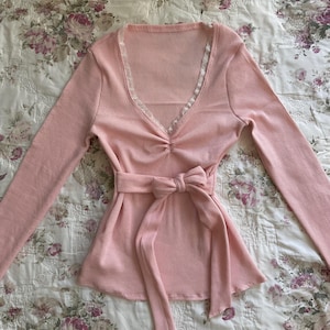 May include: A pale pink long-sleeved top with a V-neckline trimmed with white lace. The top has a gathered detail at the bust and a matching fabric belt tied in a bow at the waist. The ribbed fabric adds texture.