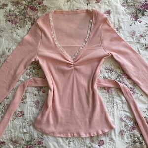 May include: A light pink long-sleeved top with a V-neckline and a tie-waist design. The top features a ribbed texture and delicate white lace trim along the neckline. The fabric appears soft and lightweight, suitable for casual wear.