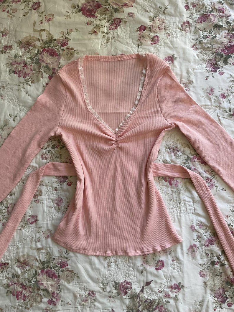 May include: A light pink long-sleeved top with a V-neckline and a tie-waist design. The top features a ribbed texture and delicate white lace trim along the neckline. The fabric appears soft and lightweight, suitable for casual wear.
