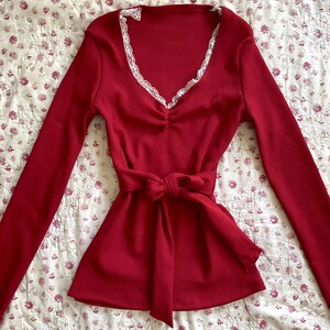 May include: A long-sleeved, red top with a V-neckline trimmed with white lace. The top has a cinched waist with a matching tie. The fabric has a ribbed texture, and the garment is displayed on a floral patterned surface.