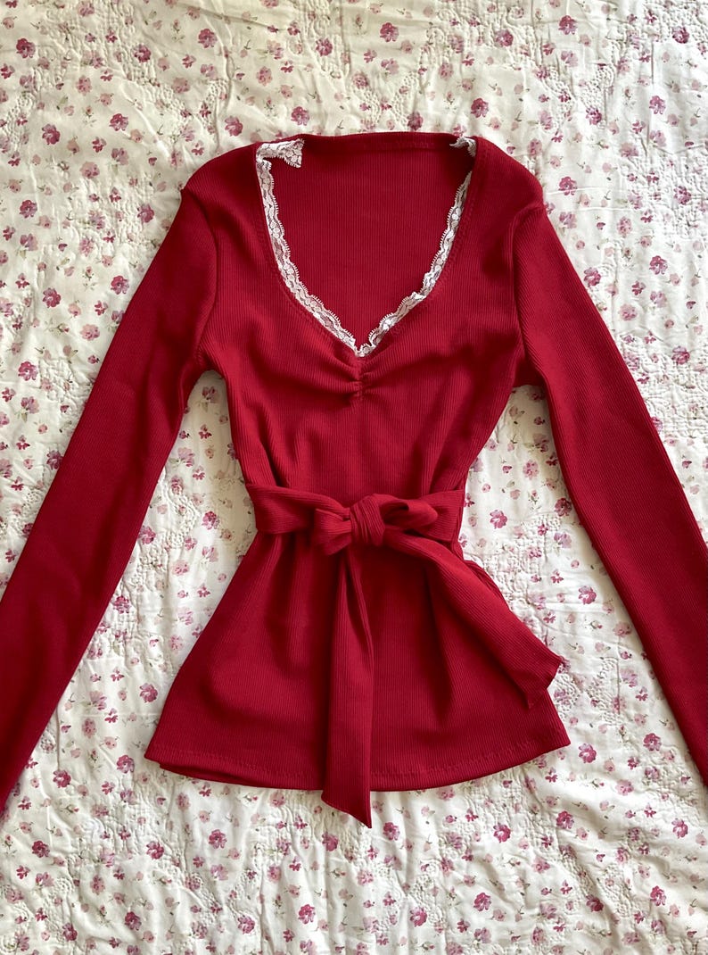 May include: A long-sleeved, red top with a V-neckline trimmed with white lace. The top has a cinched waist with a matching tie. The fabric has a ribbed texture, and the garment is displayed on a floral patterned surface.