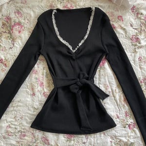 May include: Black long-sleeve top with a V-neckline trimmed with white lace. The top features a tie-waist design and is made of a ribbed fabric. The garment is displayed on a floral patterned surface.