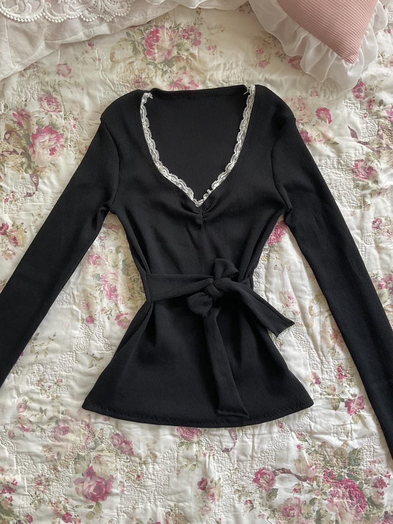 May include: Black long-sleeve top with a V-neckline trimmed with white lace. The top features a tie-waist design and is made of a ribbed fabric. The garment is displayed on a floral patterned surface.