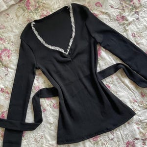 May include: Black long-sleeve top with a V-neckline trimmed with white lace. The top has a ribbed texture and a tie-waist design. The garment is displayed on a floral patterned fabric.