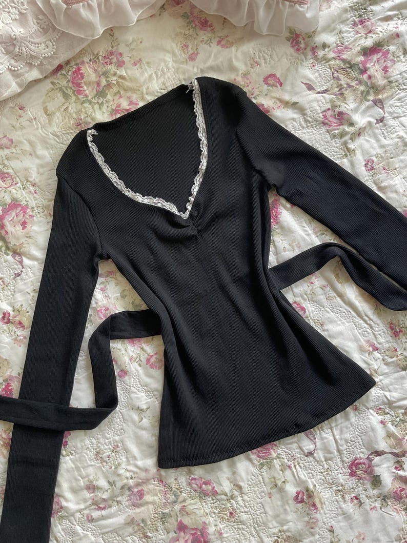 May include: Black long-sleeve top with a V-neckline trimmed with white lace. The top has a ribbed texture and a tie-waist design. The garment is displayed on a floral patterned fabric.