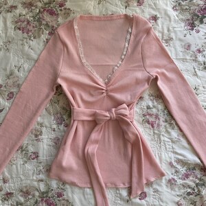 May include: A light pink long-sleeved top with a V-neckline trimmed with lace. The top features a gathered detail at the bust and a matching fabric belt tied in a bow at the waist. The ribbed fabric adds texture.