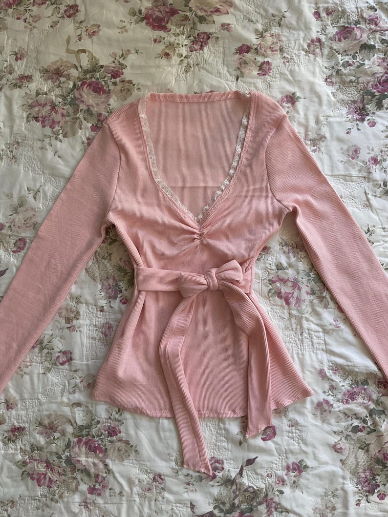 May include: A light pink long-sleeved top with a V-neckline trimmed with lace. The top features a gathered detail at the bust and a matching fabric belt tied in a bow at the waist. The ribbed fabric adds texture.