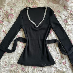 May include: A black long-sleeved top featuring a V-neckline edged with white lace. The top has a ribbed texture and a tie at the waist. The text "@missflora1111" is visible near the bottom hem.