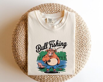 May include: A cream colored t-shirt with a cartoon bull fishing in a lake. The text "Bull Fishing" is printed in black above the bull.