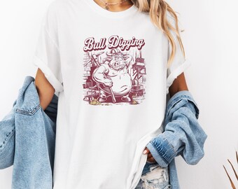 May include: White t-shirt with a red graphic of a bull wearing a hard hat and holding a pickaxe. The text "Bull Digging" is above the bull.