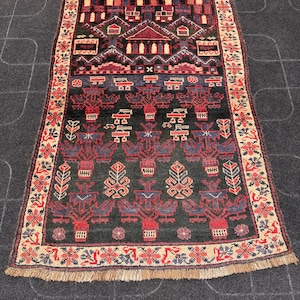 Puede incluir: Alfombra rectangular tejida a mano con un campo central verde oscuro y intrincados patrones geométricos en rojo, azul y beige. El borde presenta motivos florales y un borde con flecos. El diseño de la alfombra incluye formas estilizadas de casas y árboles.